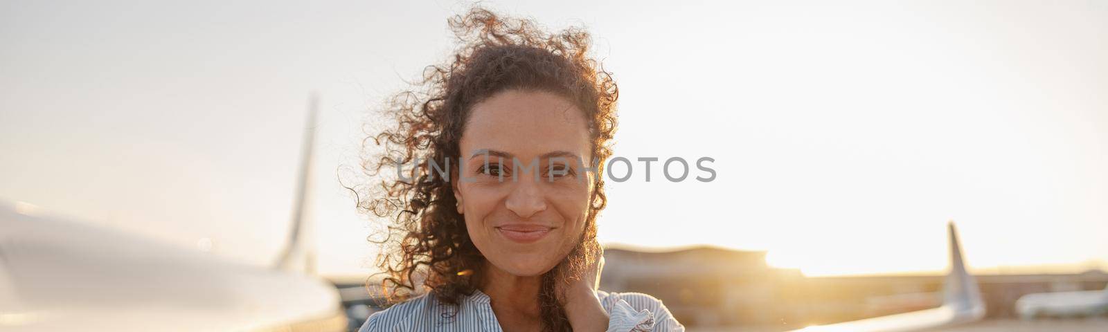 Portrait of relaxed woman smiling at camera while standing outdoors ready for boarding the plane at sunset. Vacation, lifestyle, traveling concept