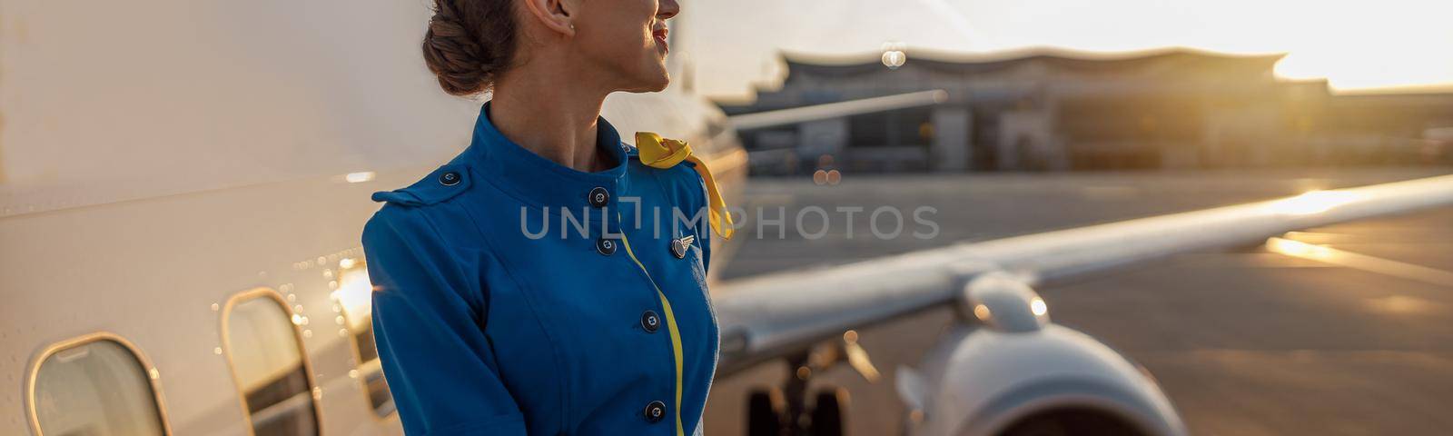 Thoughtful air stewardess in blue uniform looking away, standing outdoors at the sunset. Commercial airplane near terminal in an airport in the background by Yaroslav_astakhov