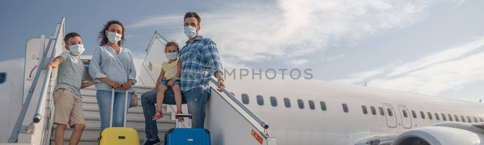 Happy family in protective masks, parents with two little children standing on airstair, boarding the plane in the daytime by Yaroslav_astakhov