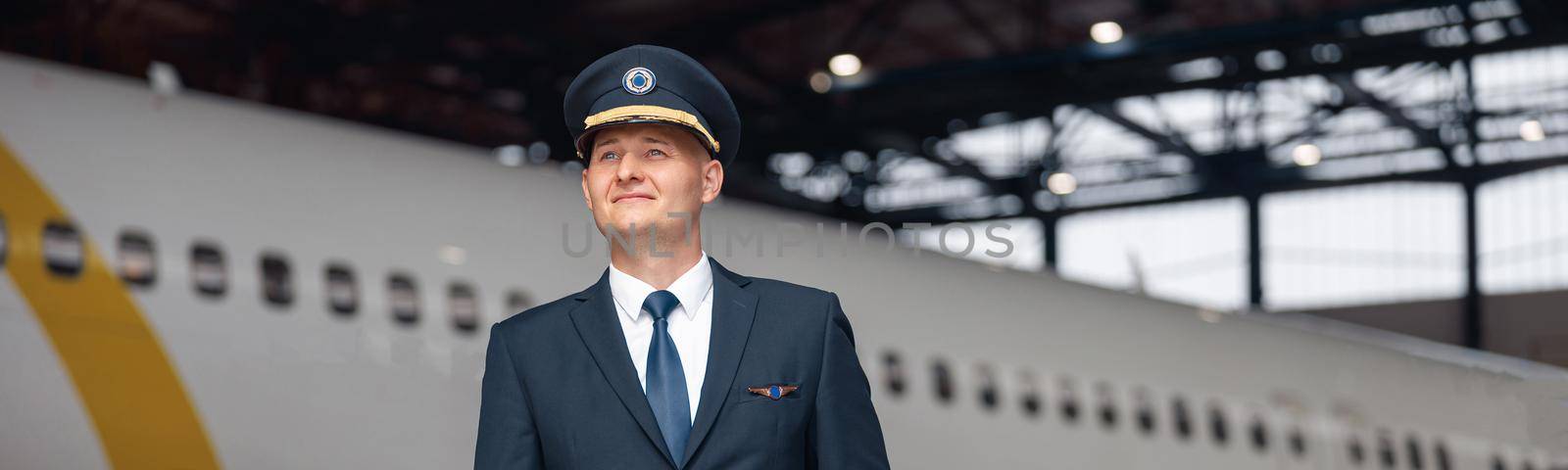 Thoughtful pilot in uniform looking away, standing in front of big passenger airplane in airport hangar. Aircraft, occupation, transportation concept