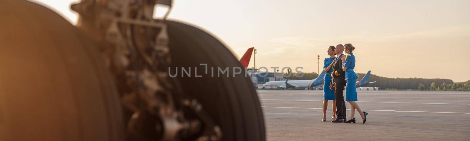 Full length shot of male pilot posing for photoshoot together with two air hostesses in blue uniform, standing in an airport terminal at sunset. Aircraft, aircrew concept