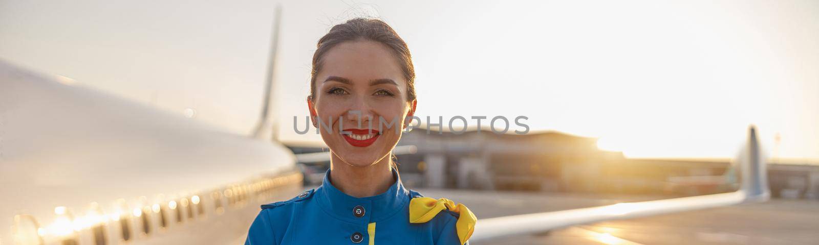 Portrait of beautiful air stewardess with red lips in blue uniform smiling at camera, posing outdoors with commercial airplane near the terminal in an airport in the background by Yaroslav_astakhov