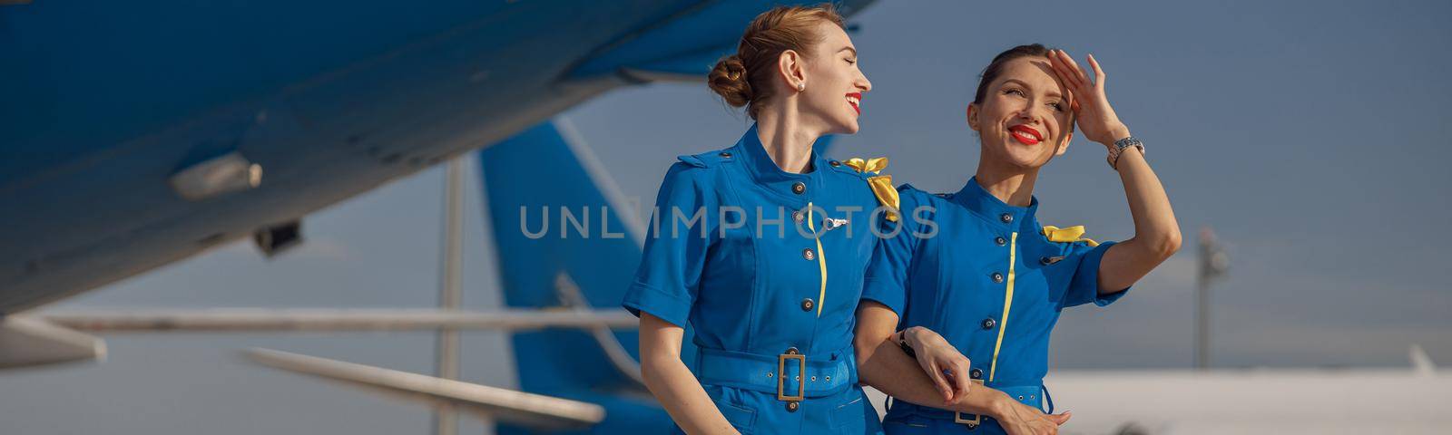 Two cheerful air stewardesses in bright blue uniform walking outdoors in front of passenger aircraft on a sunny day by Yaroslav_astakhov