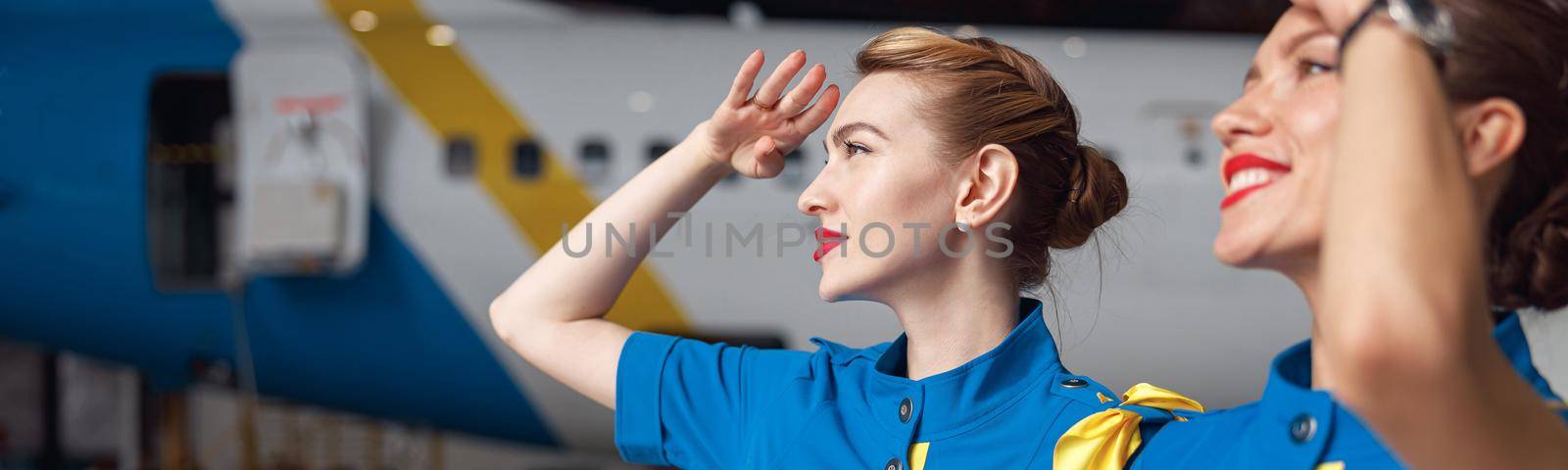 Portrait of two beautiful air stewardesses in stylish blue uniform smiling while looking up in the sky, standing together in front of passenger aircraft in hangar at the airport. Occupation concept