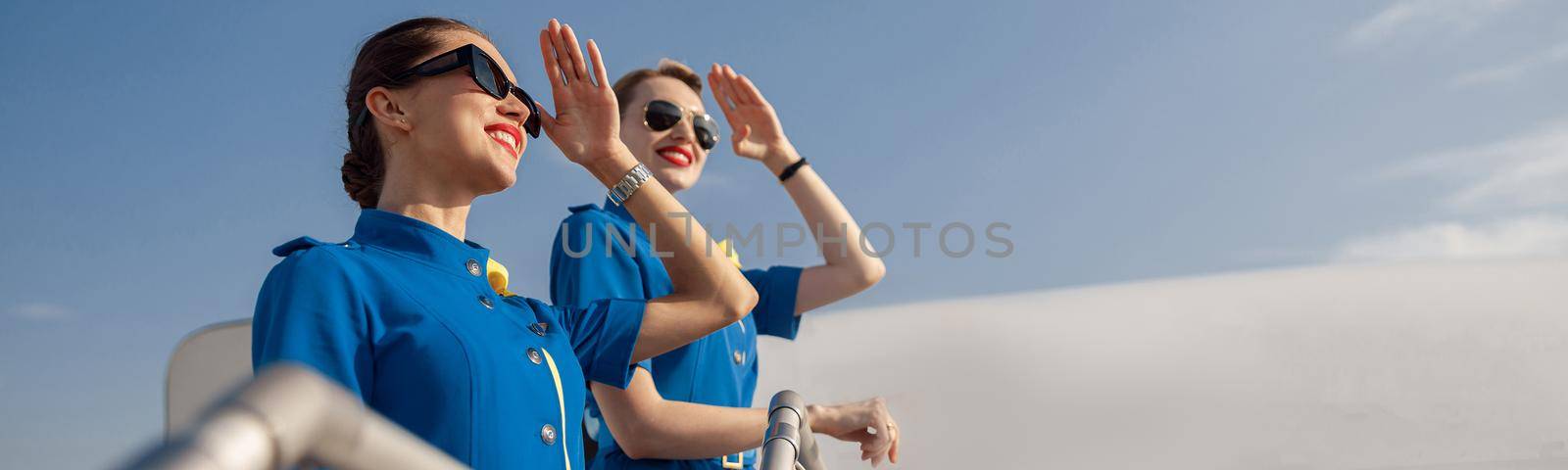 Portrait of two elegant air stewardesses in blue uniform and sunglasses covering eyes with hand and looking far away, standing together on airstair. Aircrew, occupation concept