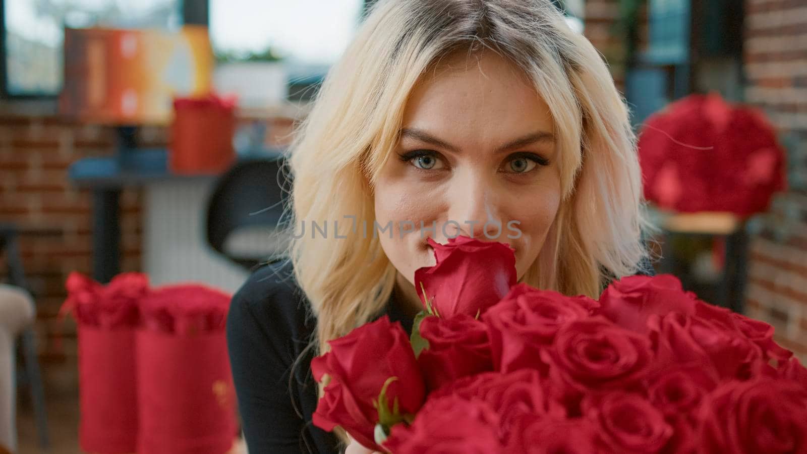 Close up of blonde attractive woman smelling bouquet with red roses enjoying surprise from boyfriend during valentine day. Lovely girlfriend surrounded by floral gift celebrating romantic anniversary