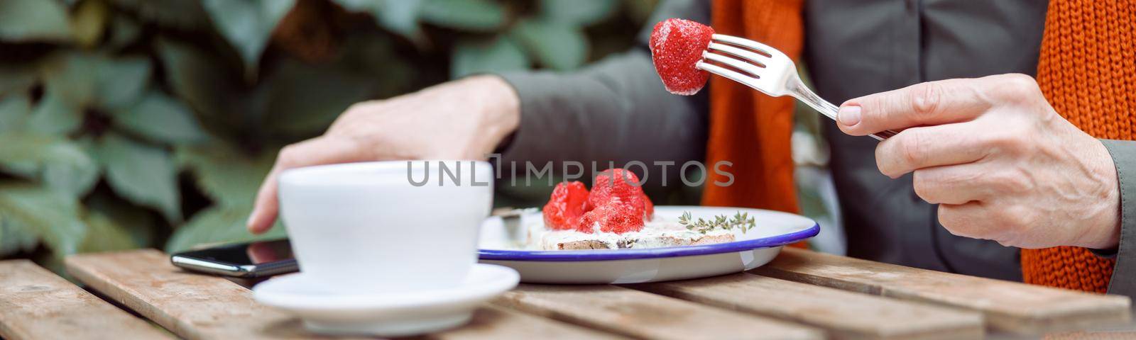 Mature woman eats delicious toast with cream and strawberries and takes mobile phone near cup at table on outdoors cafe terrace closeup