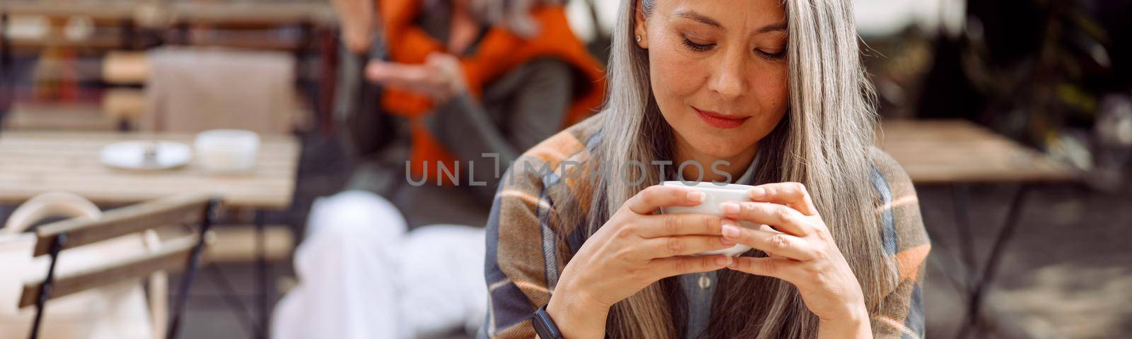 Senior cafe guests, focus on pretty silver haired lady holding cup of drink at table on outdoors cafe terrace on autumn day