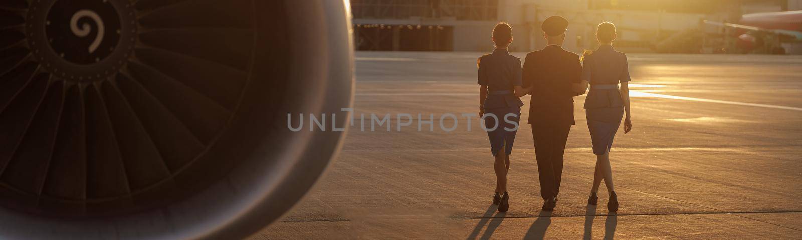 Full length shot of professional pilot walking together with two female flight attendants in blue uniform in terminal at sunset by Yaroslav_astakhov