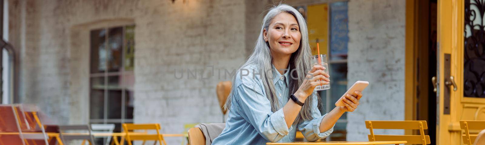 Elegant grey haired Asian woman with glass of water and smartphone sits at small table on outdoors cafe terrace