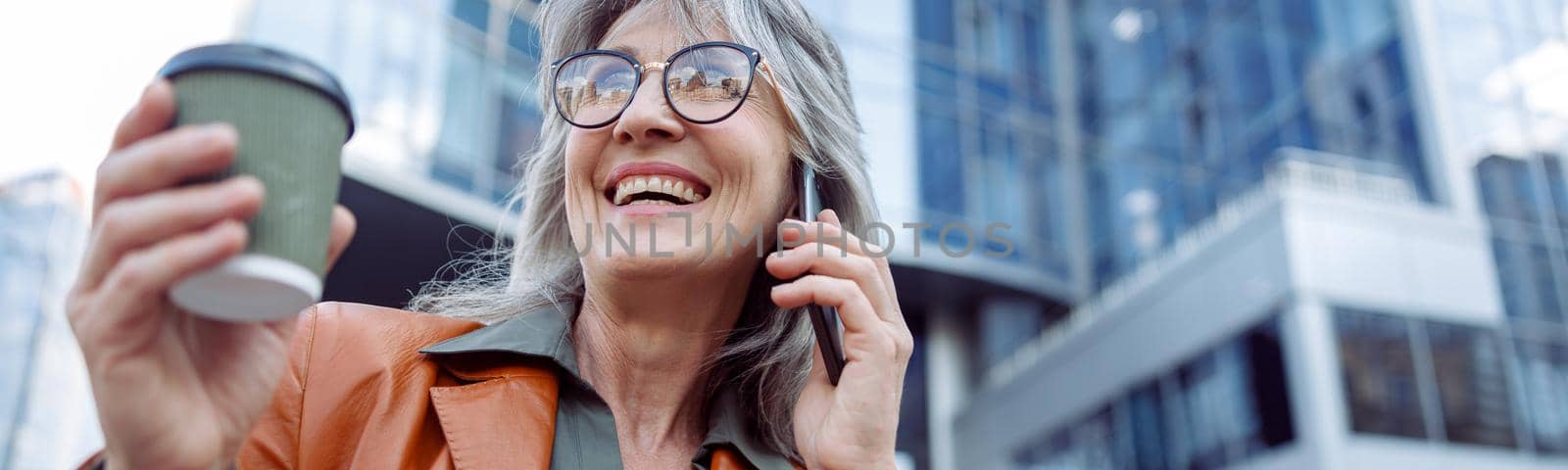 Happy female pensioner with cup of drink and glasses in talks on mobile phone on modern city street on autumn day low angle view