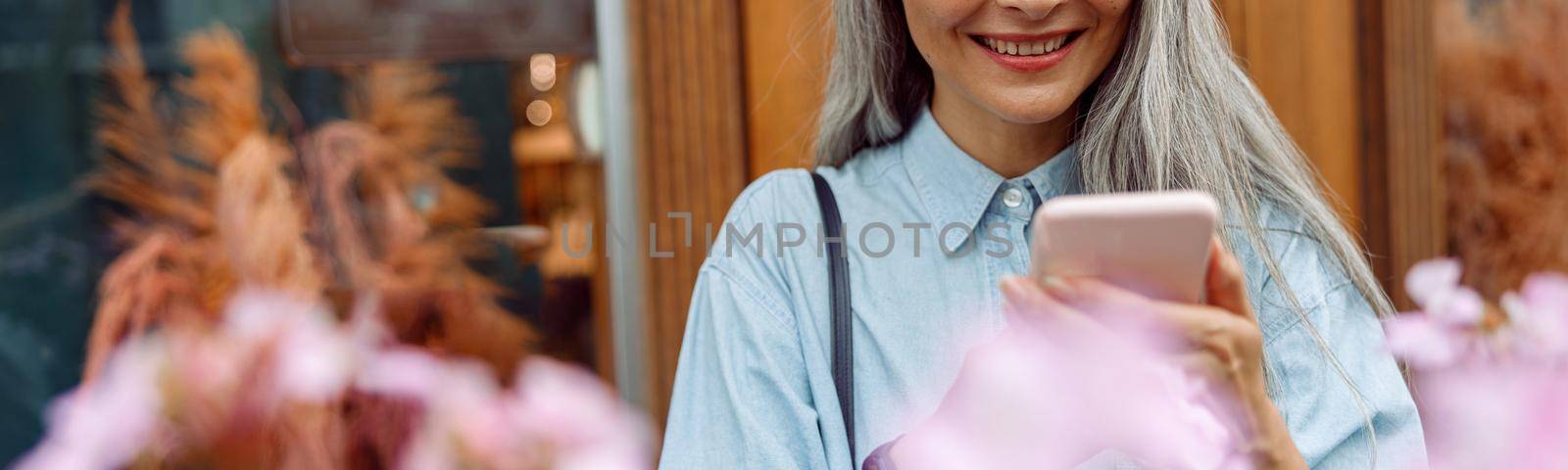 Beautiful mature Asian woman in denim shirt uses mobile phone on terrace by Yaroslav_astakhov