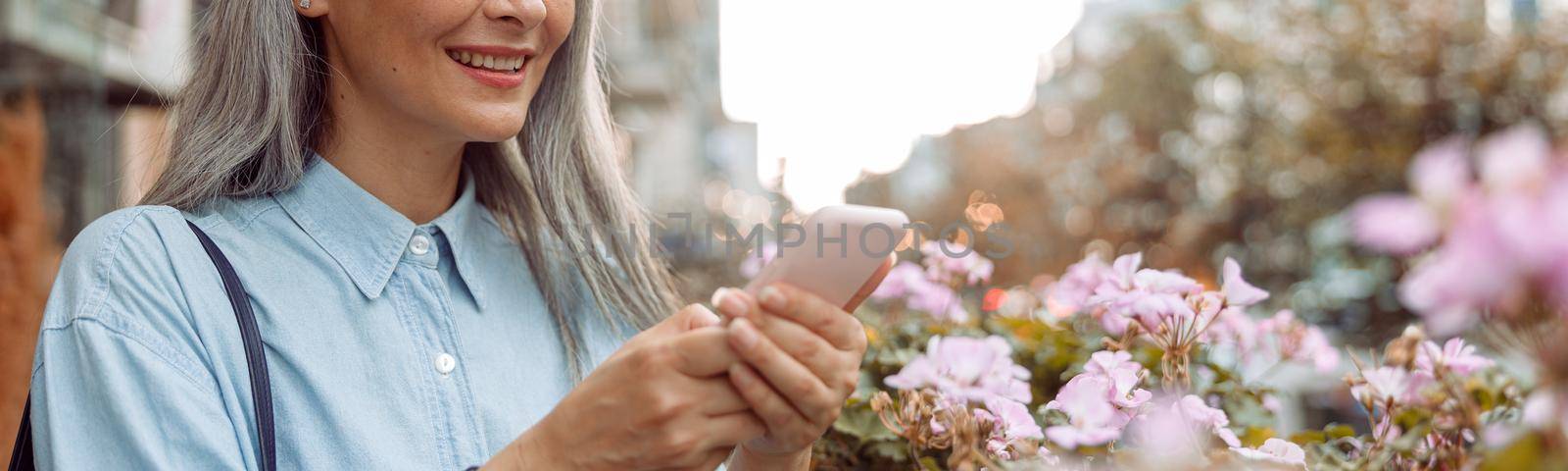 Smiling grey haired Asian woman uses cellphone standing on outdoors terrace by Yaroslav_astakhov