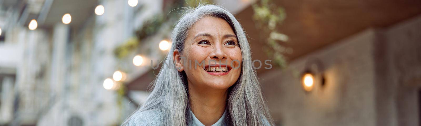 Cheerful mature Asian woman with glass of water and mobile phone sits at small table on outdoors cafe terrace on autumn day