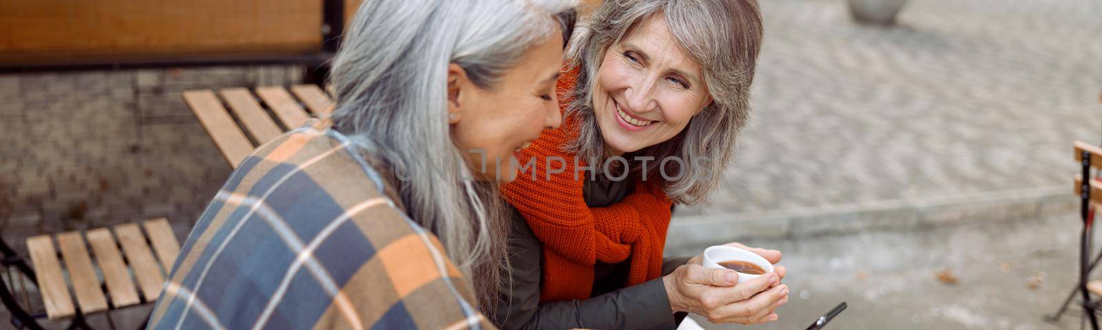 Mature Asian woman shows mobile phone to best friend in street cafe on autumn day by Yaroslav_astakhov