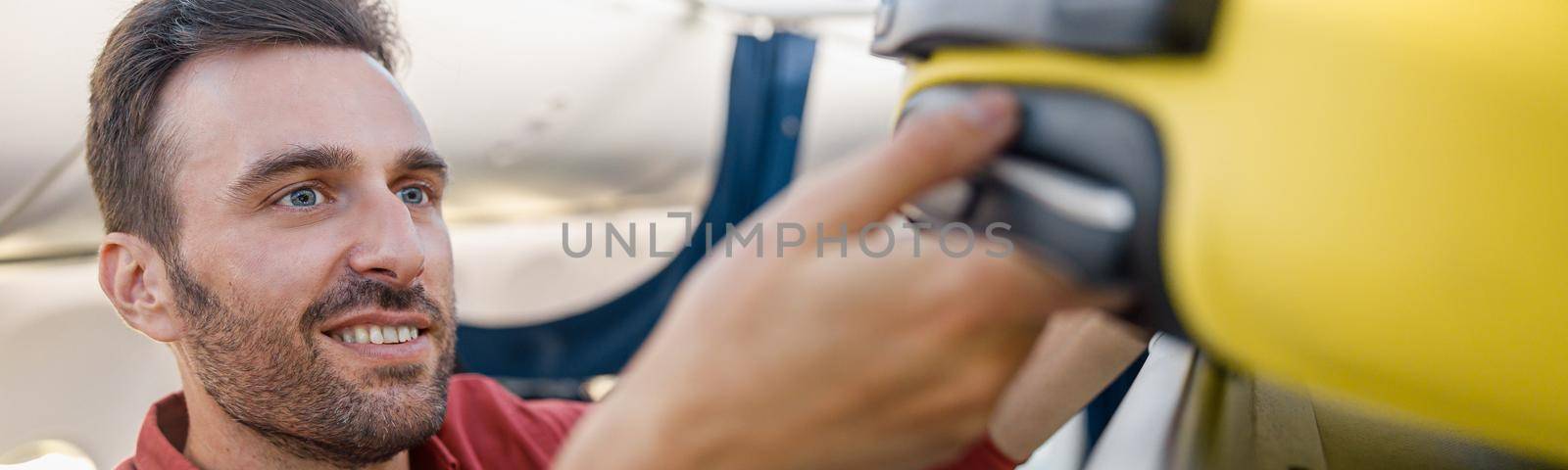 Portrait of cheerful man pulling out suitcase from hand luggage compartment, overhead locker while traveling by plane. Vacation, transportation concept