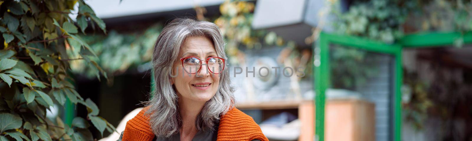 Happy silver haired mature woman with glasses eats dessert near cup of drink sitting at small table on outdoors cafe terrace on autumn day