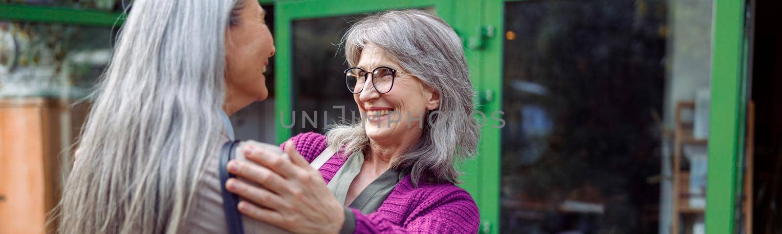 Smiling mature woman embraces female friend with long silver hair meeting on modern city street. Long-time friendship relationship