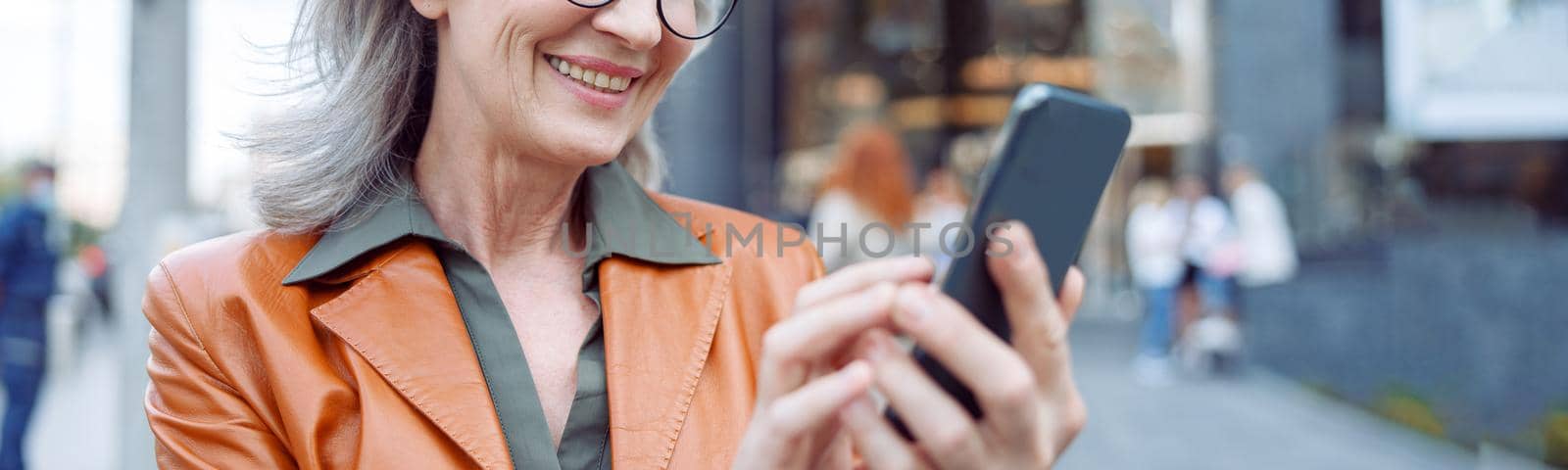 Positive hoary haired mature lady with glasses uses mobile phone standing on modern city street on autumn day