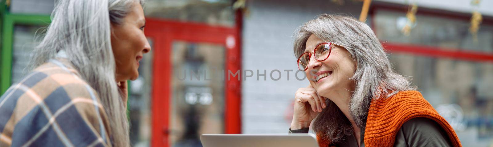 Grey haired lady with laptop spends time with Asian friend on outdoors cafe terrace by Yaroslav_astakhov