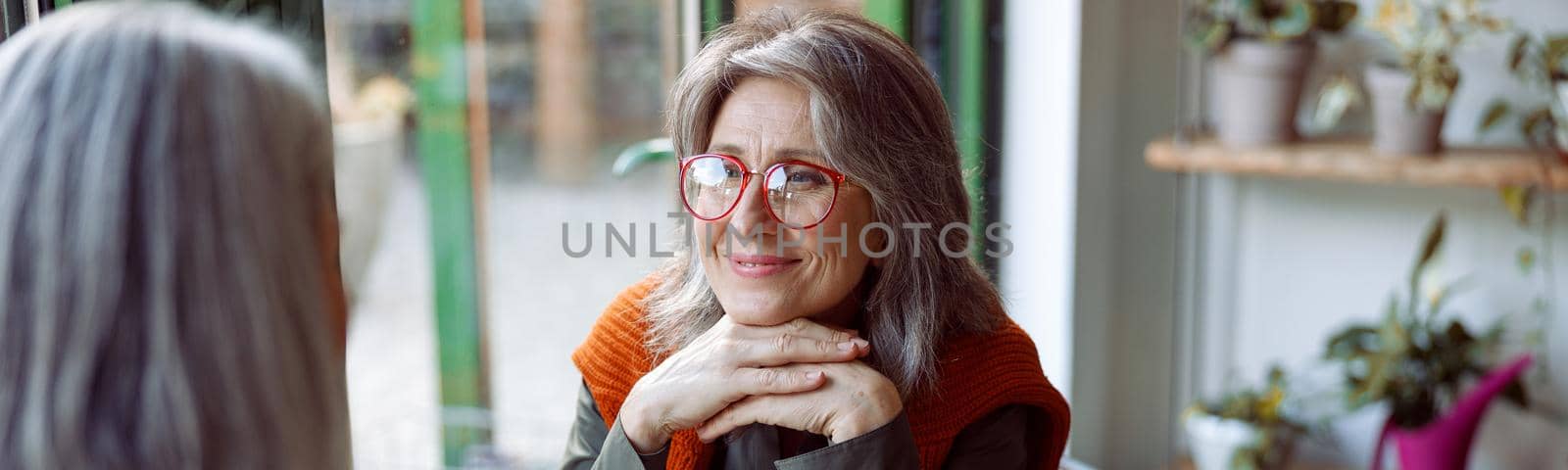 Beautiful grey haired woman with glasses looks at friend sitting at small table in cozy cafe. Long-time friendship