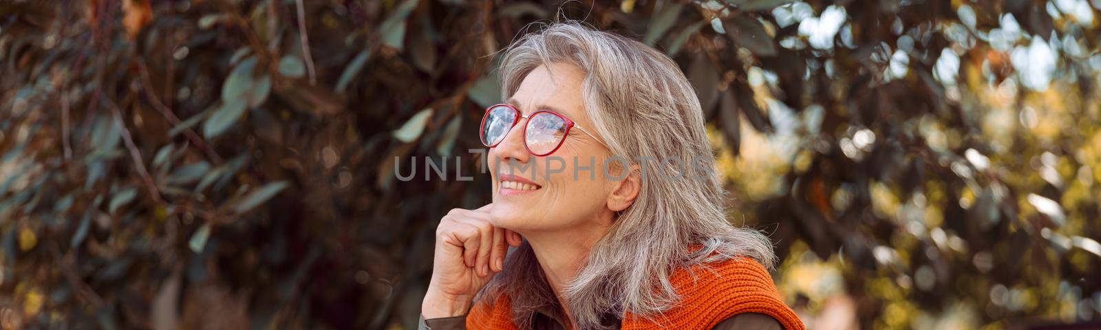 Dreamful mature lady with glasses and cup of delicious coffee sits leaning on hand at table on outdoors cafe terrace on autumn day