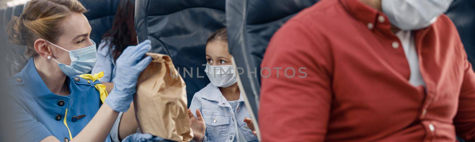 Cute girl in protective face mask looking at female flight attendant serving lunch to little passenger on board. Traveling by airplane during Covid19 pandemic by Yaroslav_astakhov
