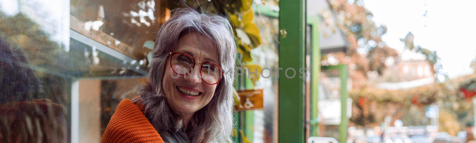 Positive mature woman with glasses and laptop holds cup of drink sitting on bench near cafe window on autumn day