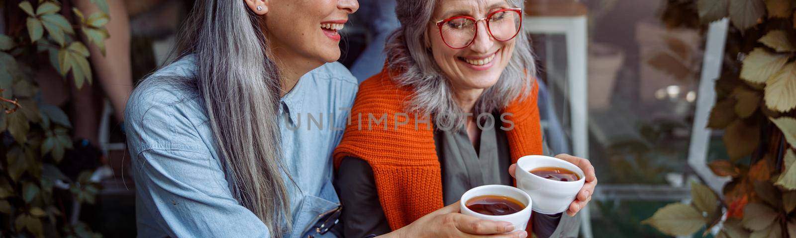 Smiling mature Asian lady with friend clink cups of coffee sitting on bench near large cafe window outdoors. Long-time friendship