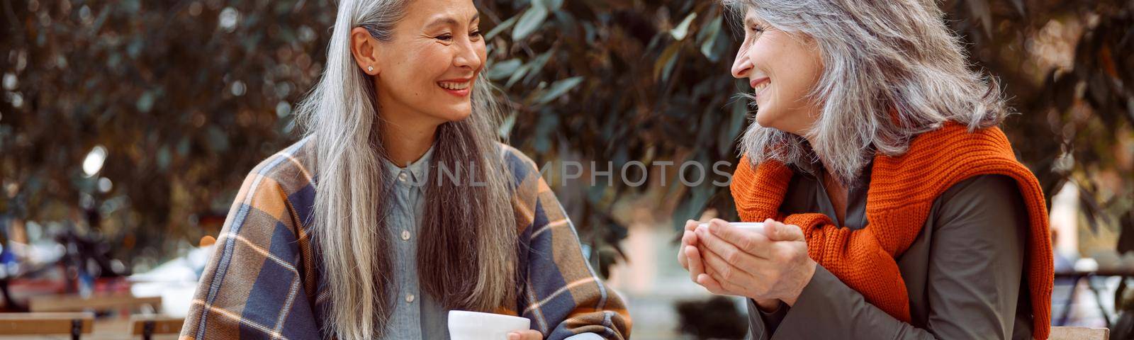 Positive mature Asian woman talks to best friend resting together at small wooden table in street cafe on nice autumn day