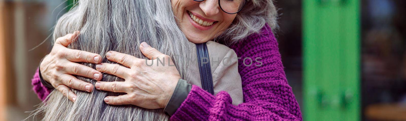 Happy senior woman with glasses in knitted jacket hugs best friend with long silver hair meeting on city street