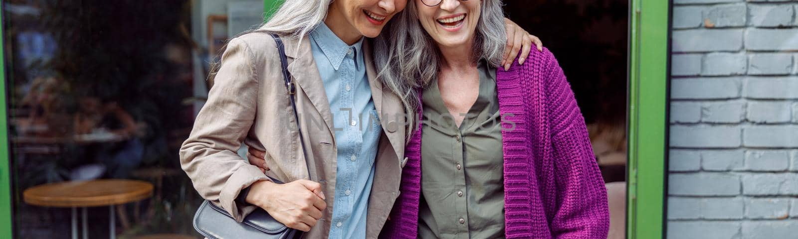 Pretty senior Asian lady hugs grey haired friend in purple jacket standing near cafe entrence on city street. Long-time friendship relationship