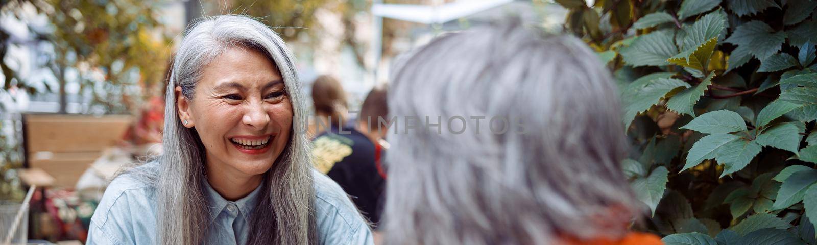 Laughing mature Asian woman with grey haired friend sit meeting at small table on outdoors cafe terrace on autumn day