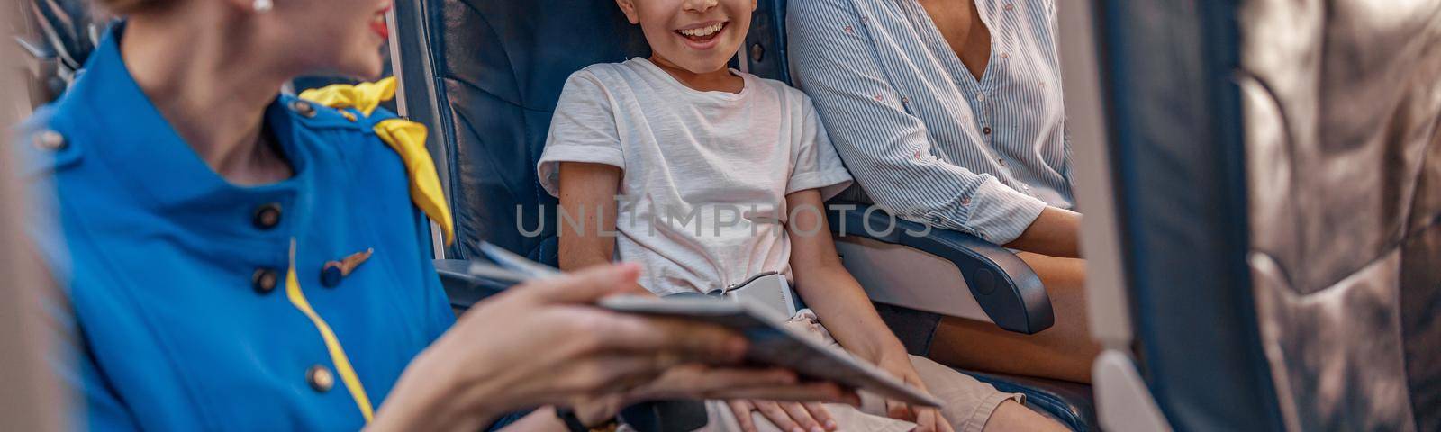 Female air hostess trying to entertain a kid on the plane by offering a book to read. Cabin crew provide service to family in airplane. Airline transportation and tourism concept