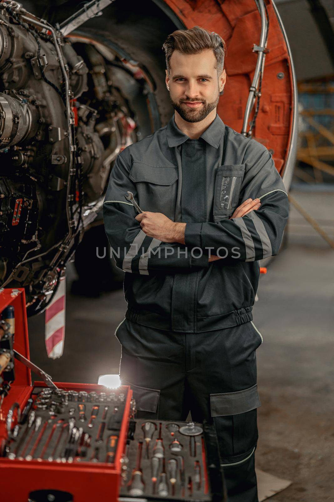 Bearded man aviation maintenance technician holding wrench and smiling while standing near airplane at repair station