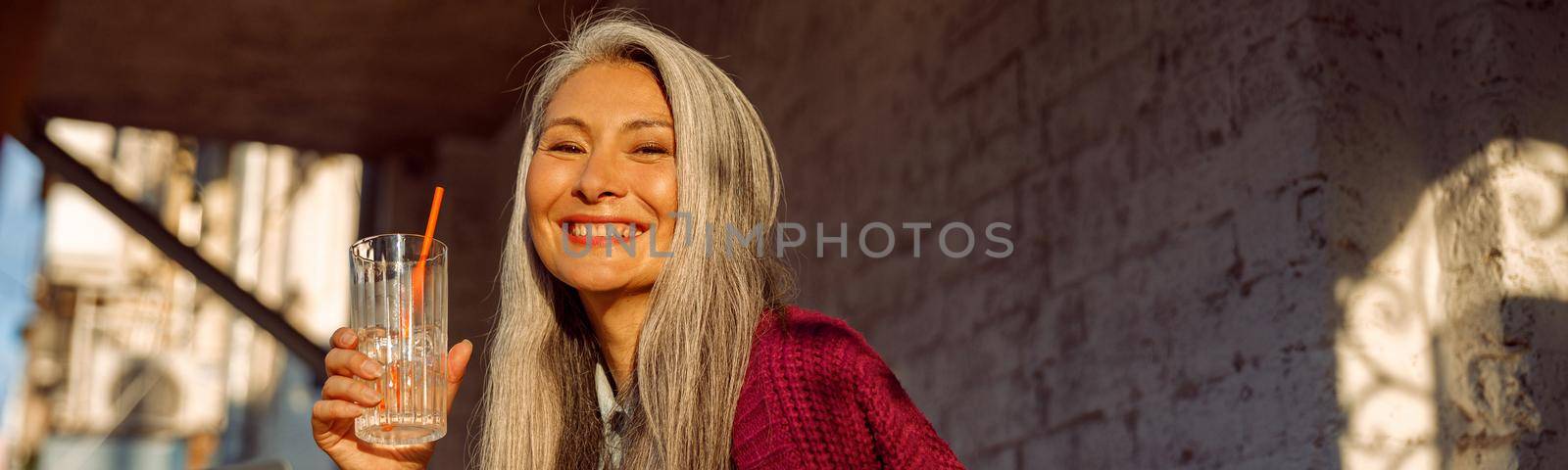 Positive mature Asian woman holds drink sitting at small table with laptop in cafe by Yaroslav_astakhov