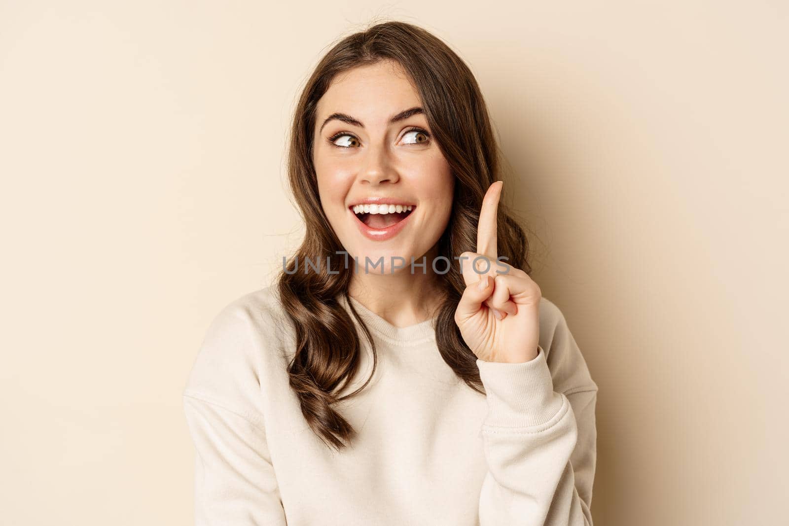 Close up shot of excited woman looking amazed with opened mouth, standing over beige background. Copy space