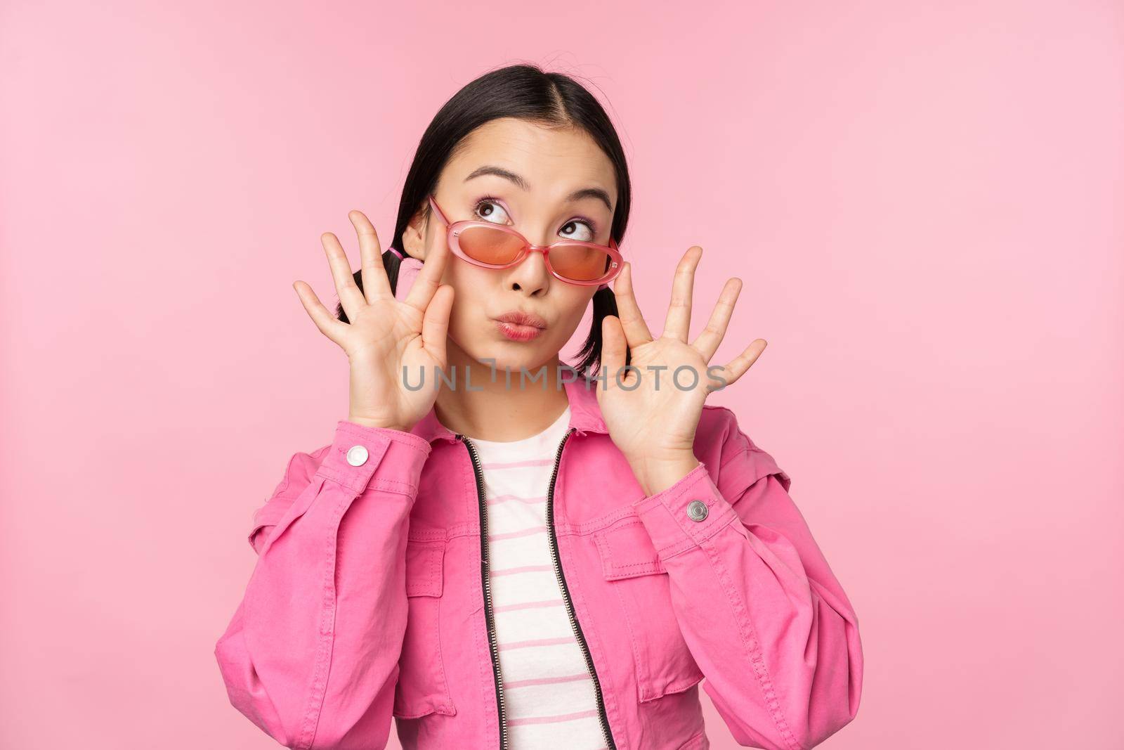 Close up of beautiful asian female model in stylish sunglasses, posing against pink background in trendy outfit, copy space by Benzoix