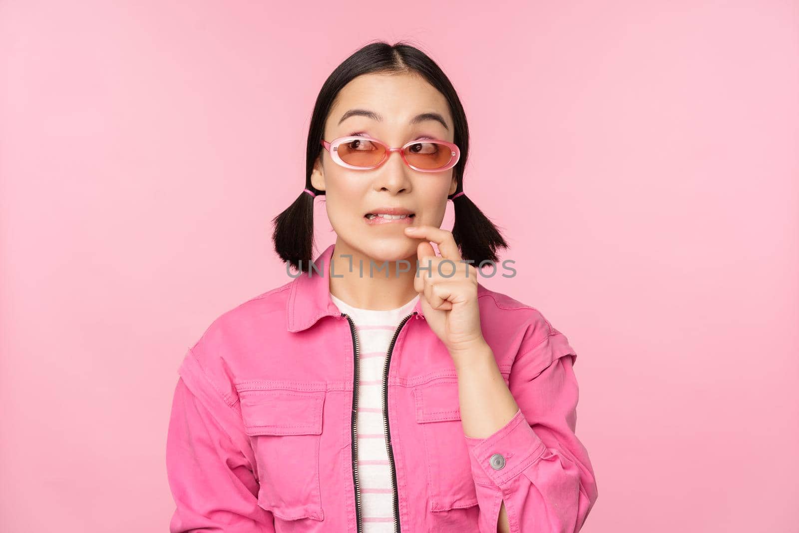 Stylish asian girl in sunglasses, looks thoughtful or intrigued, wants to try smth out, stands over pink background, pondering.