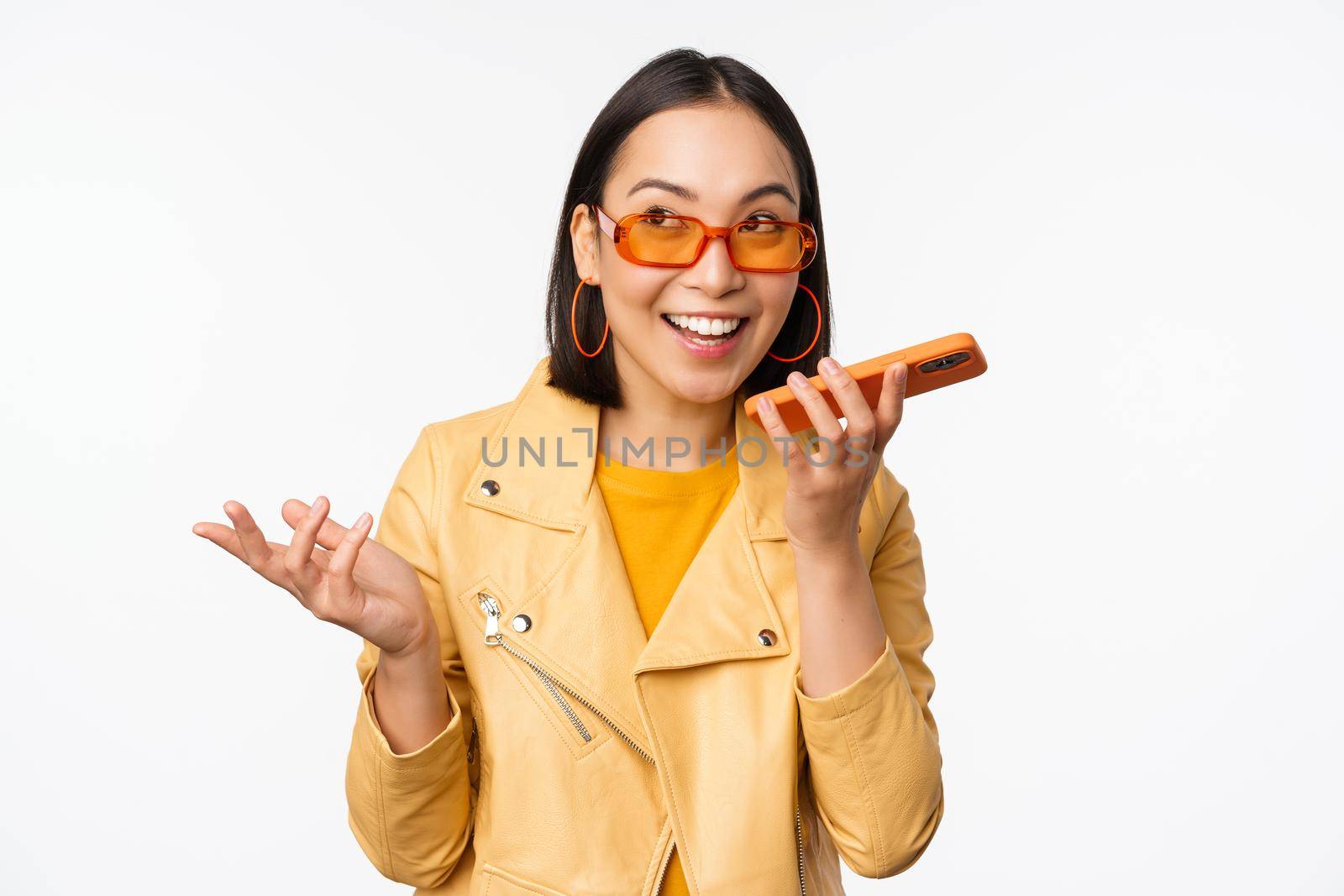 Image of happy asian girl talking on speakerphone, recording, translating her voice with mobile phone app, talking in smartphone dynamic, standing over white background by Benzoix