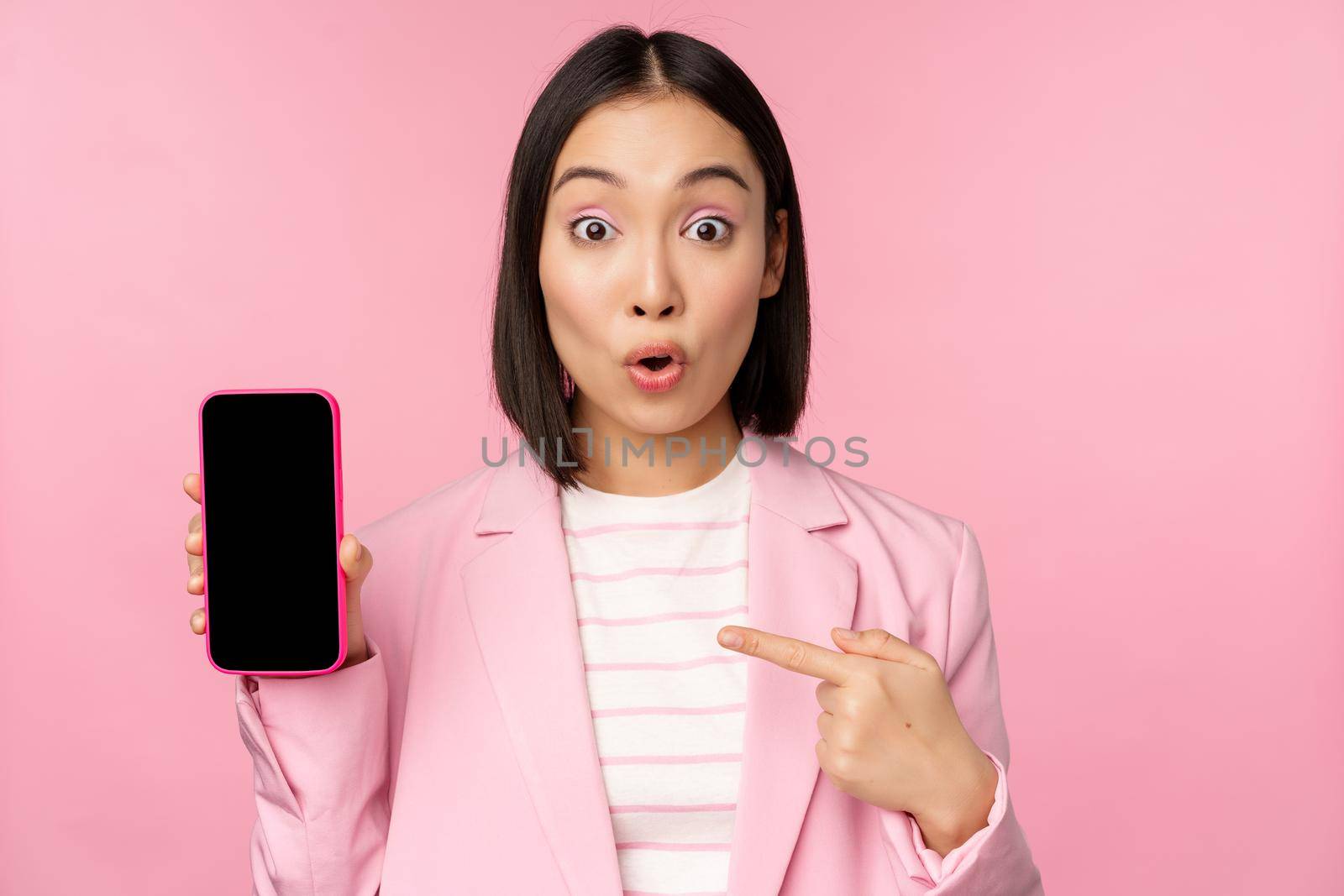 Surprised, enthusiastic asian businesswoman showing mobile phone screen, smartphone app interface, standing against pink background.