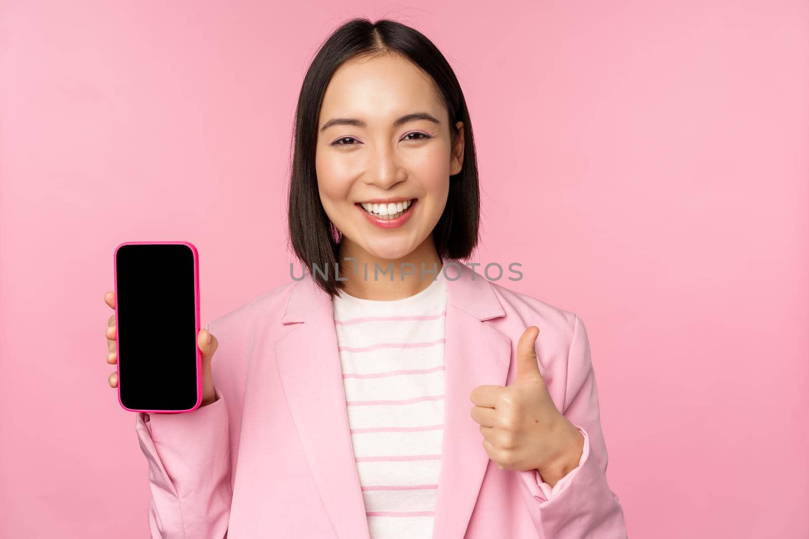 Satisfied smiling asian businesswoman recommending mobile phone app, website company on smartphone, showing screen and thumbs up, pink background by Benzoix