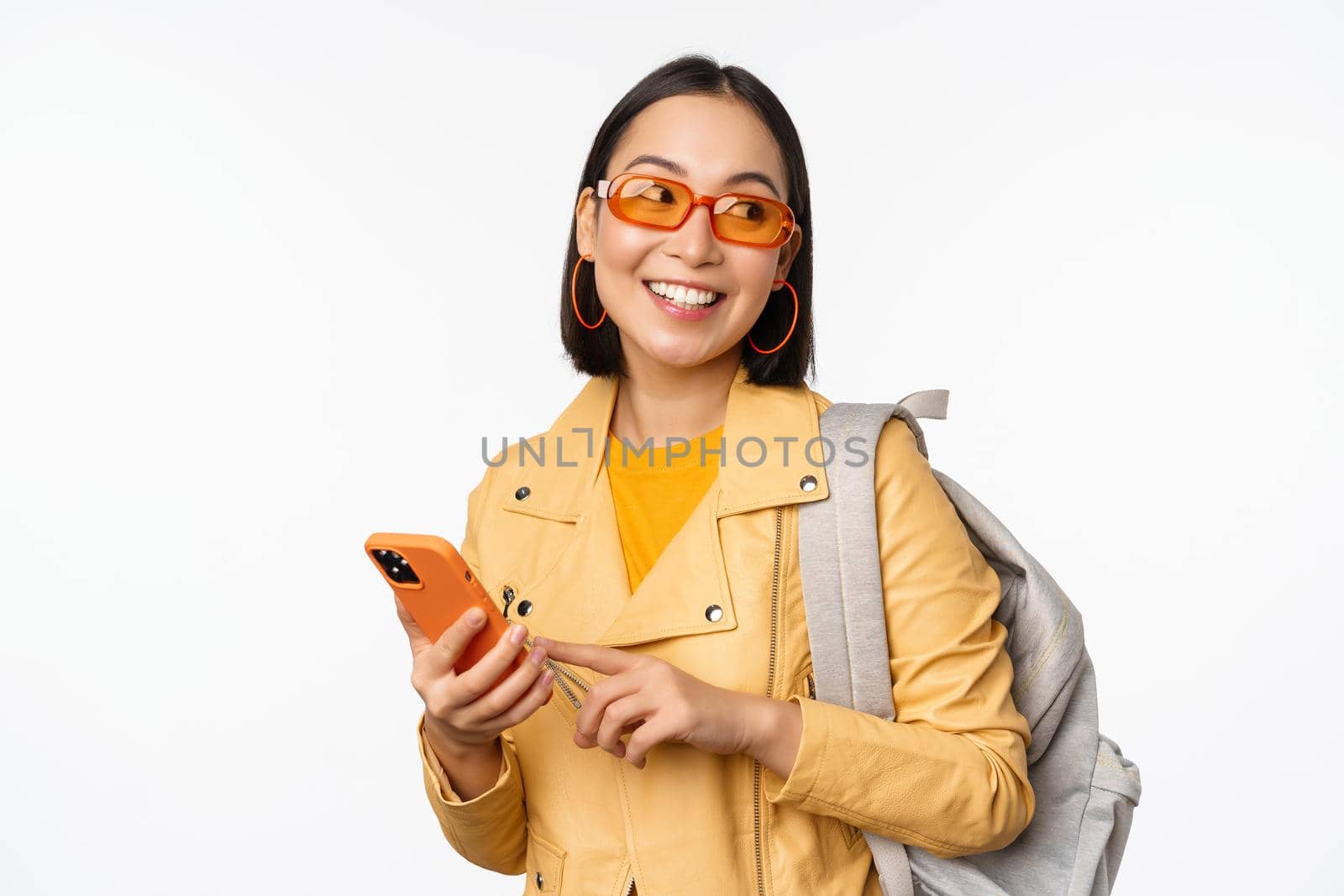 Stylish young asian woman tourist, traveller with backpack and smartphone smiling at camera, posing against white background. Copy space