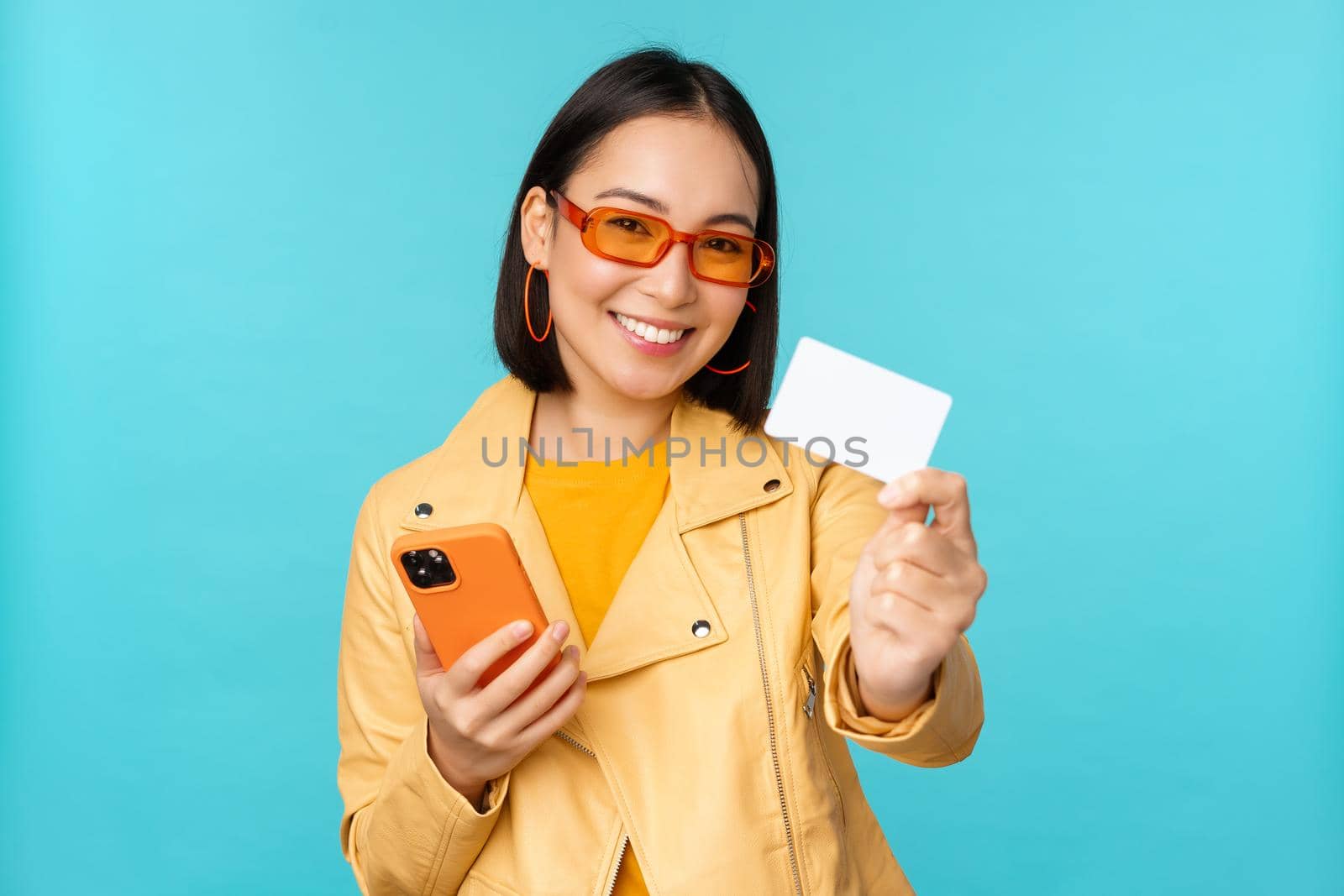Online shopping. Stylish young asian woman in sunglasses, showing credit card and using smartphone, paying in internet, making purchase, standing over blue background by Benzoix