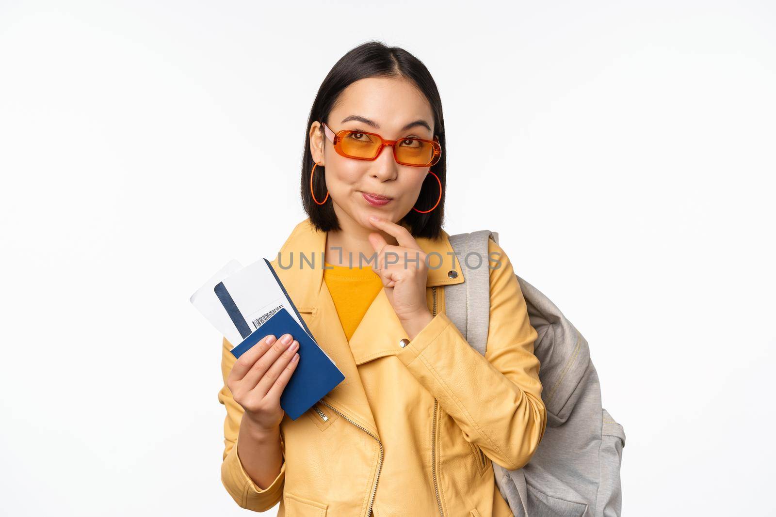 Asian girl tourist with boarding tickets and passport going abroad, holding backpack, thinking of travelling, standing over white background.