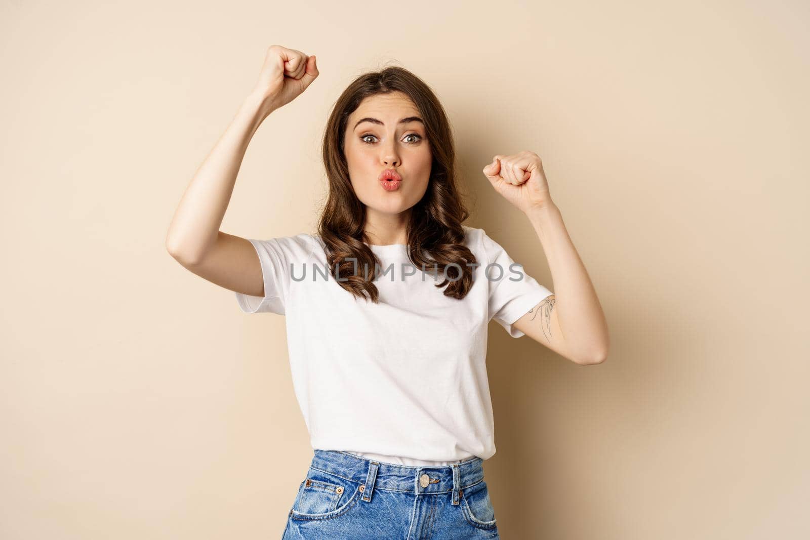 Enthusiastic adn surprised woman winning, dancing and celebrating, standing in tshirt over beige background.