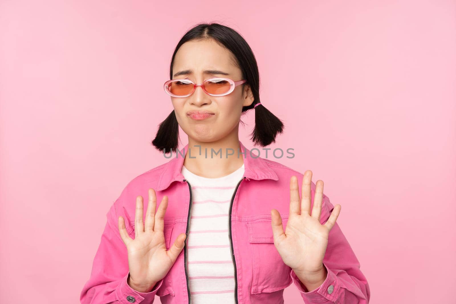 No thanks. Disgusted asian girl in sunglasses, rejecting smth, shaking hands to refuse, decline, standing over pink background.