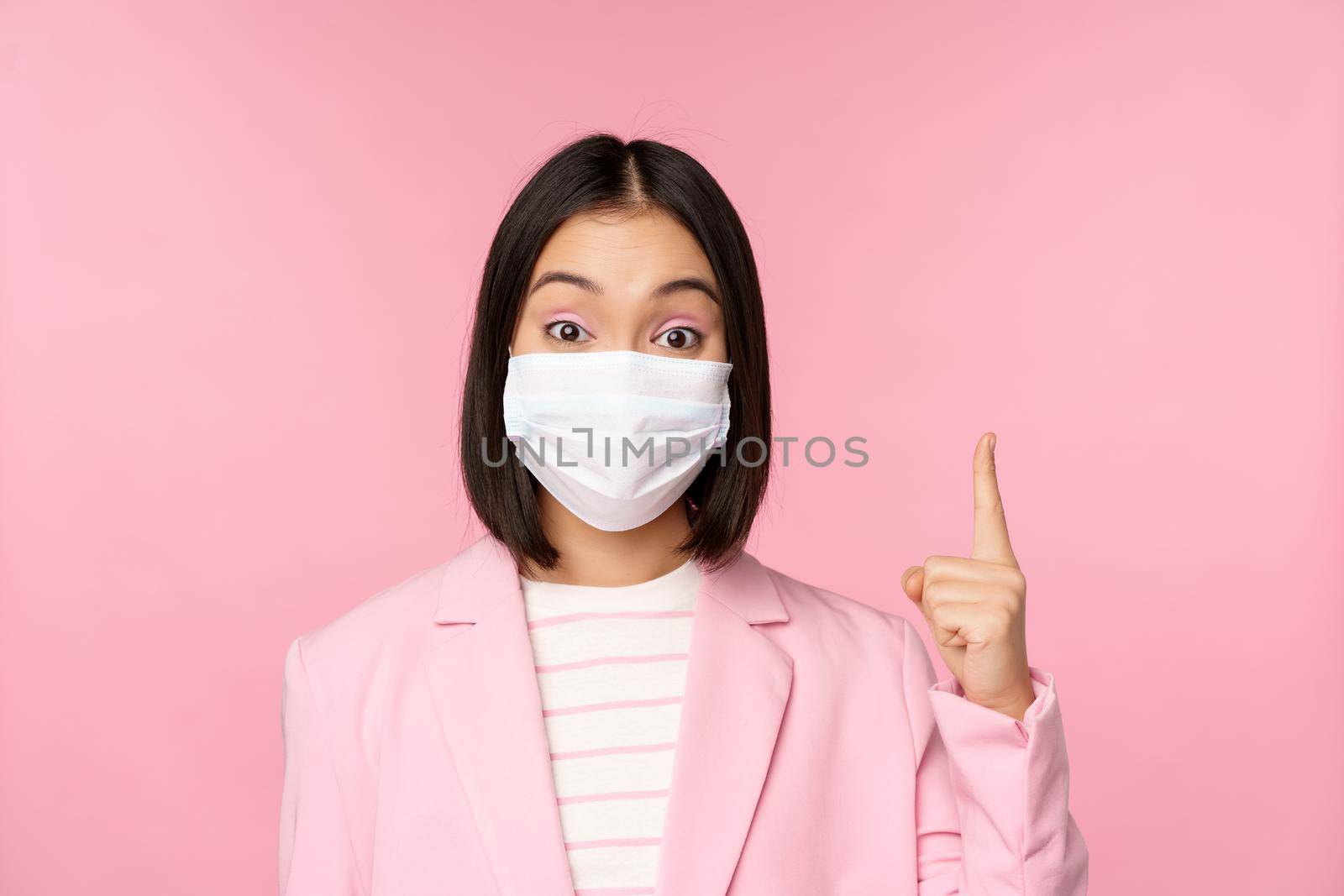 Close up portrait of asian businesswoman in medical face mask and suit, pointing finger up, showing advertisement, top banner, standing over pink background.