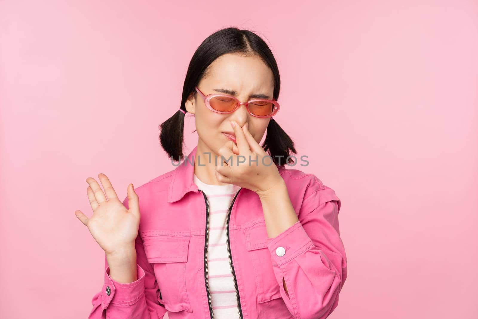 Stinky. Asian girl in sunglasses and stylish outfit, shuts her nose from disgust, dislike bad smell, standing over pink background.