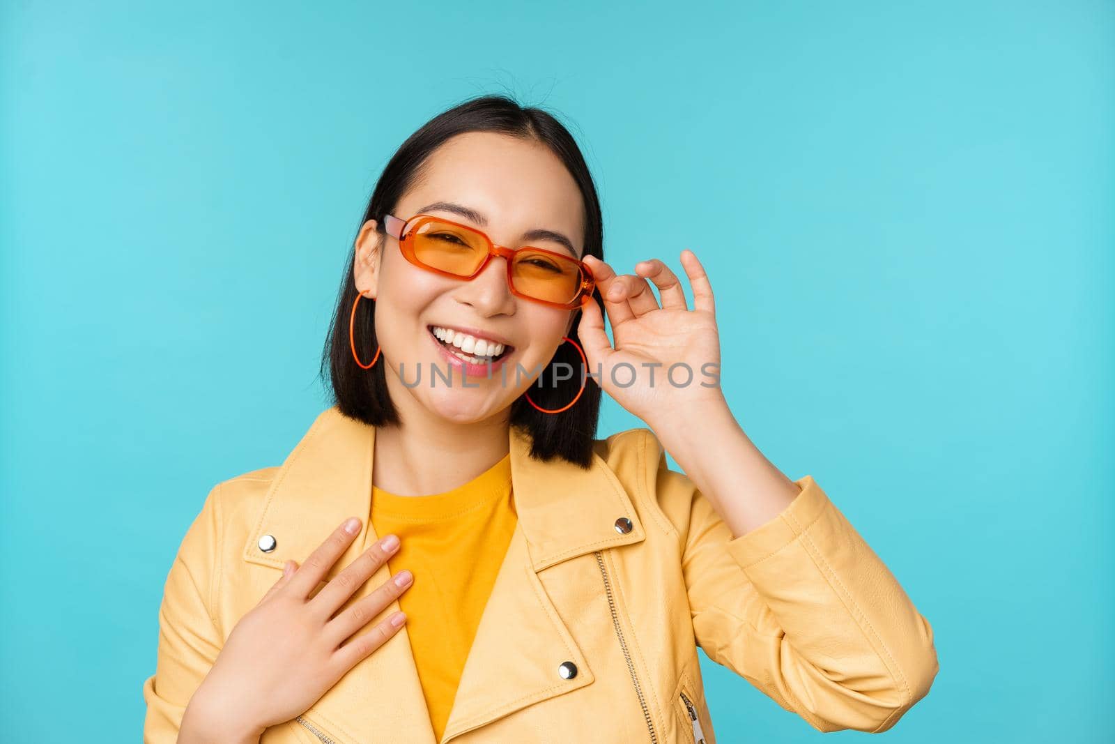 Close up portrait of stylish asian woman in sunglasses, laughing and smiling, looking happy, posing in trendy clothes over blue background by Benzoix